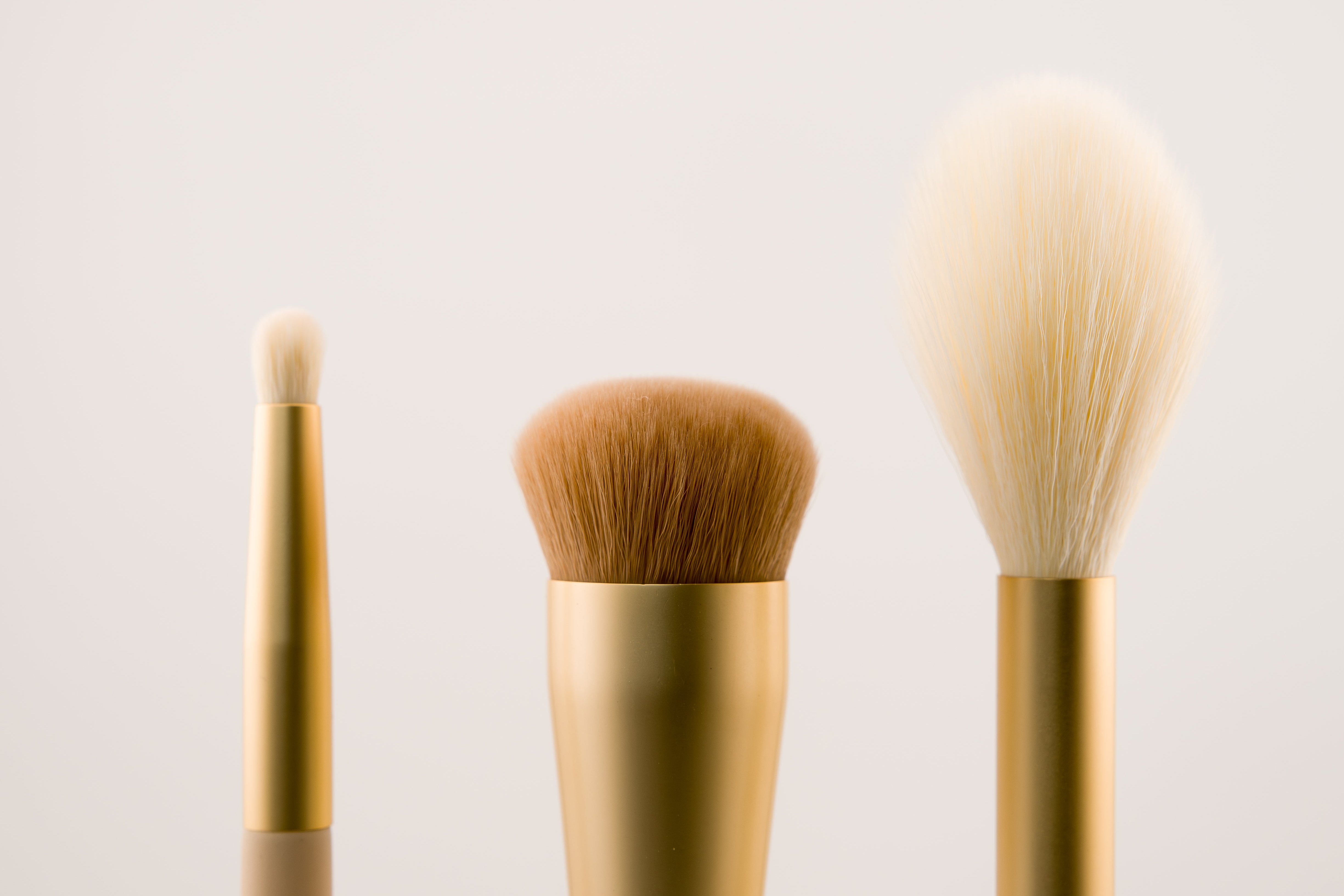 Three makeup brushes with gold handles on a light beige background